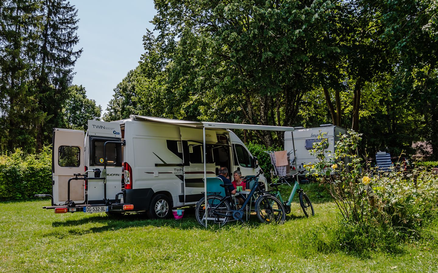Sur cette image, on voit un camping-car blanc stationné sur une pelouse verte, entouré d'arbres et de végétation. Deux p